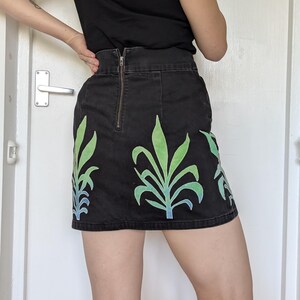 Könnte beinhalten: Schwarzer Jeans-Minirock mit hoher Taille und sichtbarem Reißverschluss. Der Rock hat ein lebendiges grün-blaues Blattdesign. Der Rock ist ein Minirock.