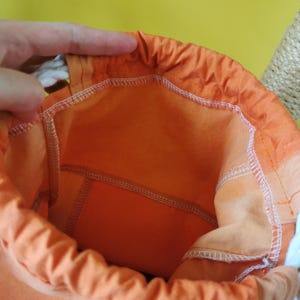 K&ouml;nnte beinhalten: Ein offener orangefarbener Beutel mit wei&szlig;em Kordelzug. Die Innenseite des Beutels ist in einem helleren Orangeton gehalten. Der Beutel besteht aus einem weichen Stoff und hat wei&szlig;e N&auml;hte.