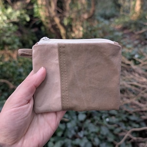 Könnte beinhalten: Eine kleine, rechteckige, zweifarbige Canvas-Geldbörse in Beige- und Brauntönen. Die Geldbörse hat einen Reißverschluss mit einem cremefarbenen Reißverschluss und eine kleine Stoffschlaufe an der Seite. Der Hintergrund ist verschwommen, mit grünem Laub.