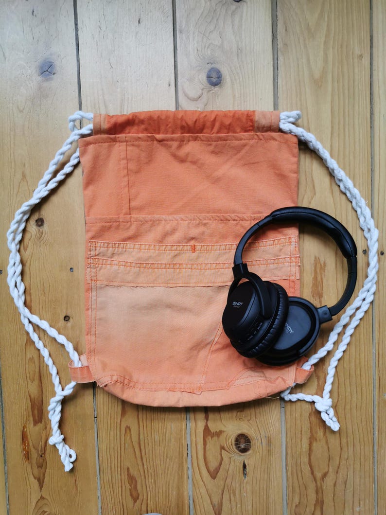 K&ouml;nnte beinhalten: Ein orangefarbener Rucksack mit wei&szlig;en Kordelz&uuml;gen und einem schwarzen Over-Ear-Kopfh&ouml;rer. Der Rucksack hat ein Taschen-Design auf der Vorderseite.