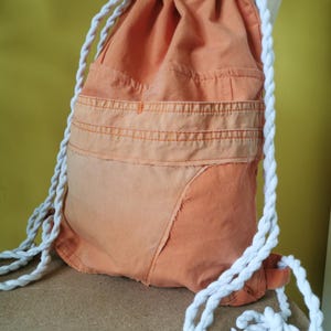 K&ouml;nnte beinhalten: Orangener Rucksack mit Kordelzug und wei&szlig;en Seilriemen. Die Tasche hat ein Patchwork-Design mit einem helleren orangefarbenen Abschnitt am Boden. Die Tasche besteht aus einem strukturierten Stoff und hat eine Tasche auf der Vorderseite.