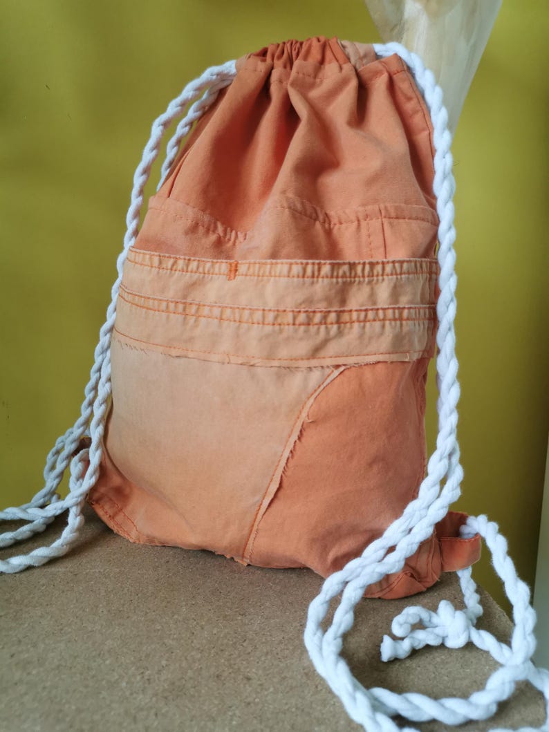 K&ouml;nnte beinhalten: Orangener Rucksack mit Kordelzug und wei&szlig;en Seilriemen. Die Tasche hat ein Patchwork-Design mit einem helleren orangefarbenen Abschnitt am Boden. Die Tasche besteht aus einem strukturierten Stoff und hat eine Tasche auf der Vorderseite.
