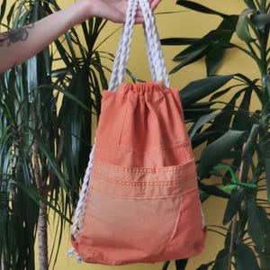 K&ouml;nnte beinhalten: Orangener Rucksack mit Kordelzug und wei&szlig;en Seilriemen. Die Tasche hat zwei Vordertaschen und einen Farbverlauf. Der Rucksack wird vor einem gelben Hintergrund mit gr&uuml;nen Pflanzen gehalten.