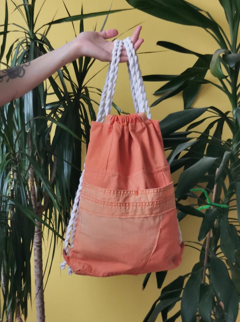 K&ouml;nnte beinhalten: Orangener Rucksack mit Kordelzug und wei&szlig;en Seilriemen. Die Tasche hat zwei Vordertaschen und einen Farbverlauf. Der Rucksack wird vor einem gelben Hintergrund mit gr&uuml;nen Pflanzen gehalten.