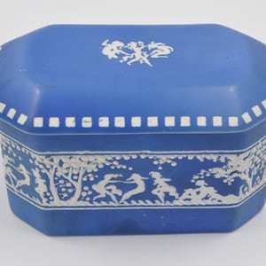 May include: A blue ceramic box with a white design of dancing figures. The box has a lid with a white border and a white design of dancing figures.