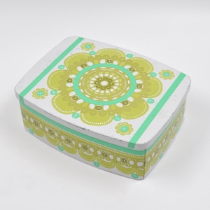 Può includere: Una scatola di latta rettangolare con base bianca, ornata da un motivo floreale nei toni del verde e del turchese. Il coperchio presenta un grande motivo floreale centrale e i lati sono decorati con un bordo smerlato. Strisce verdi accentuano i bordi.