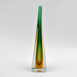 Peut inclure: Un vase en verre triangulaire et élancé, avec une base transparente et un dégradé de couleurs. Le haut est vert, passant au jaune puis à l'orange à la base. Le vase a un design élégant et moderne.
