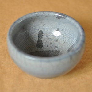 May include: A small, round, ceramic bowl with a light blue glaze. The bowl has a slightly textured surface and a dark blue interior. The bowl is likely handmade and has a rustic appearance.