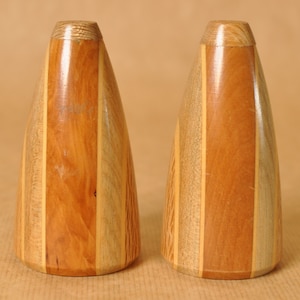 Könnte beinhalten: Zwei Salz- und Pfefferstreuer aus Holz mit konischer Form. Die Streuer bestehen aus verschiedenen Holztönen, darunter hell- und dunkelbraun und beige. Die Oberseiten der Streuer sind heller.