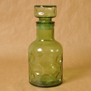 May include: A green glass bottle with a stopper. The bottle has a rounded shape with embossed circles. The stopper is square with a matching design. The bottle is likely around 20 cm tall.