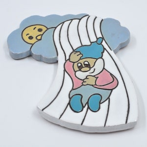 Puede incluir: Un azulejo de cerámica con una ilustración de dibujos animados de un gnomo deslizándose por un tobogán blanco y azul a rayas. El gnomo lleva un sombrero azul, una camisa rosa y pantalones azules. Un sol amarillo con cara está en el fondo.