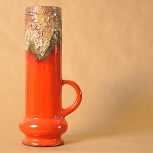 Puede incluir: Un jarrón de cerámica alto y cilíndrico en un tono naranja vibrante, con un esmalte texturizado y goteado en tonos marrones y verdes en la parte superior. El jarrón tiene un asa pequeña y una base redondeada. El jarrón mide unos 30 cm de alto.