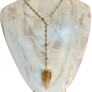 Puede incluir: Un collar dorado con un colgante de cristal de citrino. El collar presenta una delicada cadena con pequeñas cuentas cuadradas y un acento circular. El cristal de citrino es de color ámbar claro y está engastado en un marco dorado.