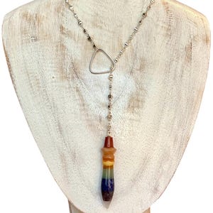 May include: A silver-toned lariat necklace featuring a delicate chain with small metallic beads and a triangular link. The pendant is a multi-colored, layered gemstone pendulum with horizontal bands of red, orange, yellow, green, blue, and purple.