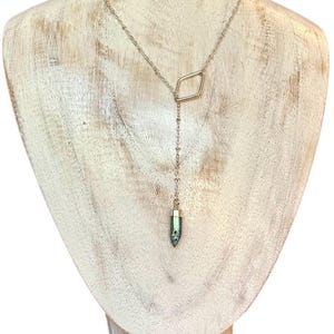 May include: A silver necklace featuring a diamond-shaped pendant and a bullet-shaped, iridescent stone. The necklace is displayed on a wooden bust. The pendant is connected to a delicate chain. The stone is a pearlescent colour.