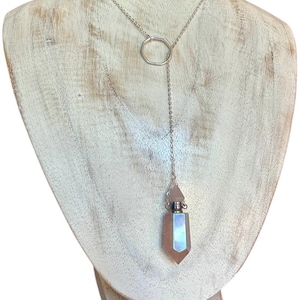 May include: A silver-toned necklace with a geometric pendant. The necklace features a delicate chain that connects to a circular element, with a longer chain suspending a faceted, iridescent, pink-toned crystal bottle. The necklace is displayed on a white wooden bust.