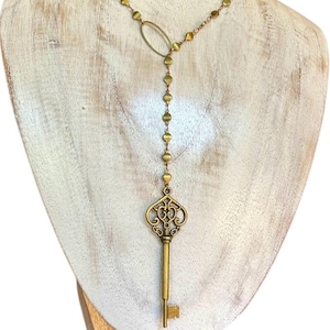 Puede incluir: Un collar dorado con un colgante de llave. El collar presenta una cadena de pequeñas cuentas redondas y un conector ovalado. El colgante de llave tiene un diseño ornamental de filigrana. El collar se exhibe en un busto de madera blanca.