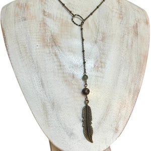 May include: A bronze-toned necklace with a feather pendant. The necklace features a delicate chain, an oval-shaped connector, and a series of small beads. The feather pendant is detailed with etched lines, adding texture to the design. The necklace is displayed on a white mannequin.