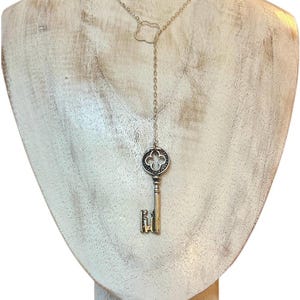 Puede incluir: Un collar plateado con un colgante de llave. La llave tiene una cabeza decorativa y un paletón detallado. El collar tiene una cadena delicada con un adorno en forma de nube cerca del cierre. El colgante cuelga de una cadena más larga.