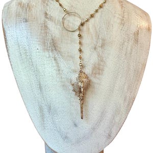 May include: A gold-toned necklace with a seashell pendant. The necklace features a circular accent and a chain of small beads. The seashell is a light tan color with gold accents, and is displayed on a white mannequin.