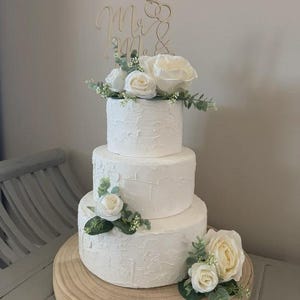 May include: A three-tiered white wedding cake with textured icing, decorated with white roses and foliage. The cake sits on a wooden stand. A gold-coloured "Mr. & Mrs." cake topper is placed on top.