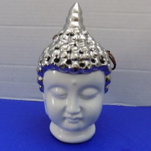 Puede incluir: Maceta de cerámica blanca en forma de cabeza de Buda con parte superior metálica plateada. La maceta tiene una expresión de ojos cerrados y un pequeño agujero en la parte superior para el drenaje.