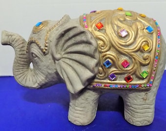 May include: A gray elephant figurine with a gold-colored blanket adorned with colorful, square-shaped jewels. The elephant has a raised trunk and detailed ears. The figurine is a decorative item.