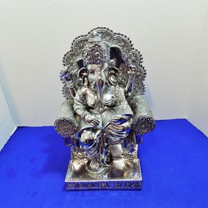 Puede incluir: Estatua de color plateado de Ganesha, el dios hind&uacute; de la sabidur&iacute;a y la prosperidad, sentado en un trono con intrincados detalles. La estatua sostiene un libro y tiene una trompa con un solo colmillo.