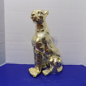 May include: A gold-colored ceramic figurine of a leopard sitting with its head turned to the side. The figurine has a spotted pattern.