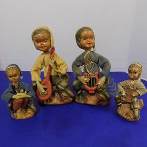Puede incluir: Conjunto de cuatro figuritas de elfos vintage, cada una tocando un instrumento musical diferente. Los elfos están vestidos con ropa colorida y tienen caras pintadas. Están sentados sobre pequeñas bases de madera.