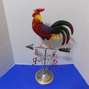 May include: A metal rooster weather vane with a red, yellow, and blue painted design. The rooster is perched on a metal stand with a compass rose and arrows pointing north, south, east, and west.