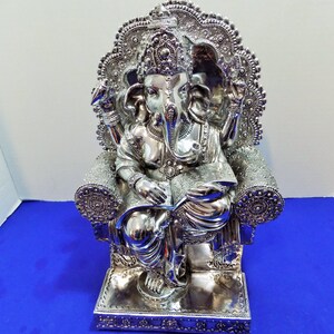 Puede incluir: Estatua plateada de Ganesha, el dios hind&uacute; de la sabidur&iacute;a y la prosperidad, sentado en un trono con un libro en su regazo. La estatua est&aacute; intrincadamente detallada con dise&ntilde;os ornamentados y una corona en la cabeza de Ganesha.