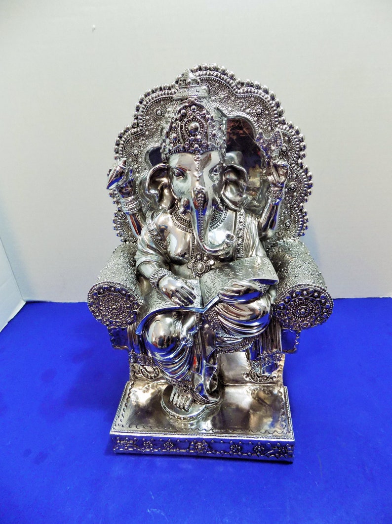 Puede incluir: Estatua plateada de Ganesha, el dios hind&uacute; de la sabidur&iacute;a y la prosperidad, sentado en un trono con un libro en su regazo. La estatua est&aacute; intrincadamente detallada con dise&ntilde;os ornamentados y una corona en la cabeza de Ganesha.