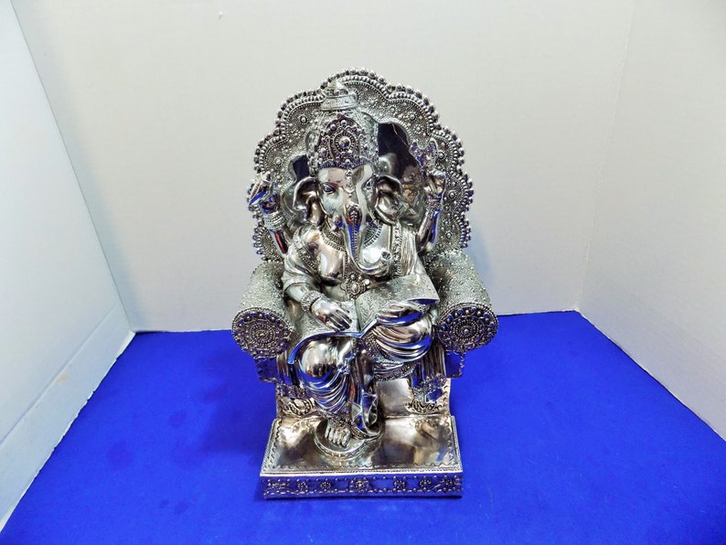 Puede incluir: Estatua de color plateado de Ganesha, el dios hind&uacute; de la sabidur&iacute;a y la prosperidad, sentado en un trono con intrincados detalles. La estatua sostiene un libro y tiene una trompa con un solo colmillo.