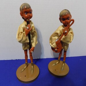 Puede incluir: Dos figuras vintage de músicos. Cada figura tiene una tez oscura, viste un atuendo de satén dorado y un anillo de perlas. Uno sostiene una flauta, el otro un laúd. Se paran sobre bases circulares marrones.