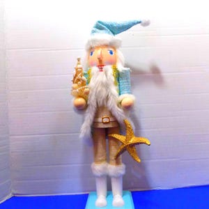 May include: A decorative nutcracker figurine in a coastal theme. It features a light blue hat and jacket, a white beard, and holds a golden tree. A golden starfish is attached to the front. The figurine is approximately 12 inches tall.