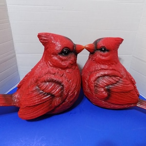 Puede incluir: Dos figuras de pájaros cardenales rojos, con ojos negros y detalles de plumas. Las aves están colocadas pico a pico. Estas figuras probablemente están hechas de un material duradero, ideal para la decoración del hogar.