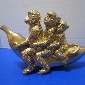 May include: A gold-colored decorative figurine featuring three monkeys sitting in a row on top of a large banana. The monkeys have textured fur details and are positioned one behind the other, with the banana forming the base of the sculpture.