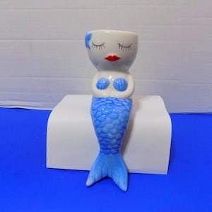 Puede incluir: Una maceta de cerámica con forma de sirena que presenta una cabeza blanca con ojos cerrados, pestañas largas y labios rojos. La sirena tiene detalles azules en forma de concha en el pecho y una cola azul texturizada con aleta.