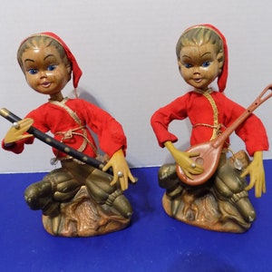Puede incluir: Dos figuras de músicos vintage. Cada figura lleva una túnica y gorro rojos, con una cara de madera. Una sostiene una flauta negra, la otra un laúd marrón. Las figuras están sentadas sobre una base dorada y marrón. Miden unos 15 cm de alto.