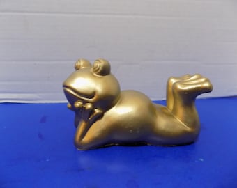 May include: A gold-colored ceramic frog figurine lying on its belly with its arms crossed and its legs extended behind it.