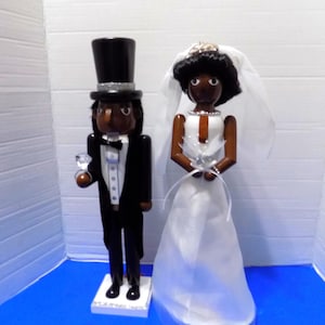 May include: Wooden nutcracker figurines of a bride and groom. The groom wears a black tuxedo with a top hat and holds a glass. The bride wears a white wedding dress and veil. The figurines are on a white base.