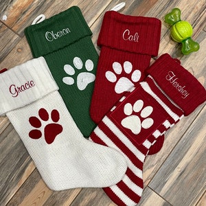 May include: Four personalized Christmas stockings with a paw print design. The stockings are red, white, and green with the names "Gracie", "Oberon", "Cali", and "Hershey" embroidered on them.