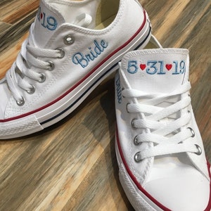 K&ouml;nnte beinhalten: Ein Paar wei&szlig;e Canvas-Sneaker mit rotem Besatz. Ein Schuh hat das Wort "Bride" in blauer Farbe gestickt. Der andere Schuh hat das Datum "5.31.19" in blauer Farbe gestickt mit zwei roten Herzen.