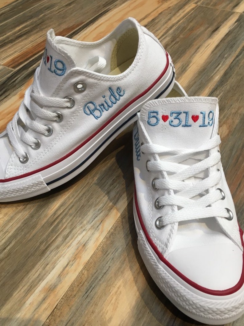 K&ouml;nnte beinhalten: Ein Paar wei&szlig;e Canvas-Sneaker mit rotem Besatz. Ein Schuh hat das Wort "Bride" in blauer Farbe gestickt. Der andere Schuh hat das Datum "5.31.19" in blauer Farbe gestickt mit zwei roten Herzen.