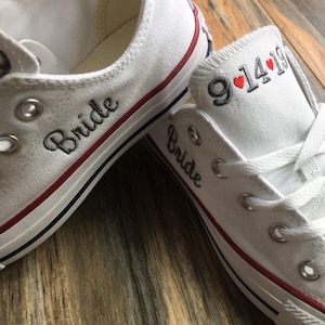 K&ouml;nnte beinhalten: Ein Paar wei&szlig;e Canvas-Schuhe mit rotem Besatz. Ein Schuh hat das Wort "Bride" auf der Seite gestickt, der andere Schuh hat das Datum "9.14.19" auf der Seite gestickt mit zwei roten Herzen.