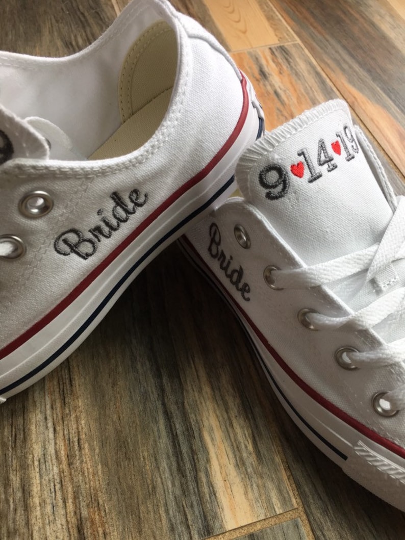 K&ouml;nnte beinhalten: Ein Paar wei&szlig;e Canvas-Schuhe mit rotem Besatz. Ein Schuh hat das Wort "Bride" auf der Seite gestickt, der andere Schuh hat das Datum "9.14.19" auf der Seite gestickt mit zwei roten Herzen.