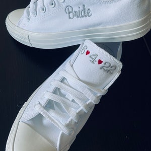K&ouml;nnte beinhalten: Ein Paar wei&szlig;e Canvas-Sneaker mit dem Wort "Bride" auf einem Schuh und dem Datum "6.4.22" mit roten Herzen auf dem anderen Schuh bestickt.