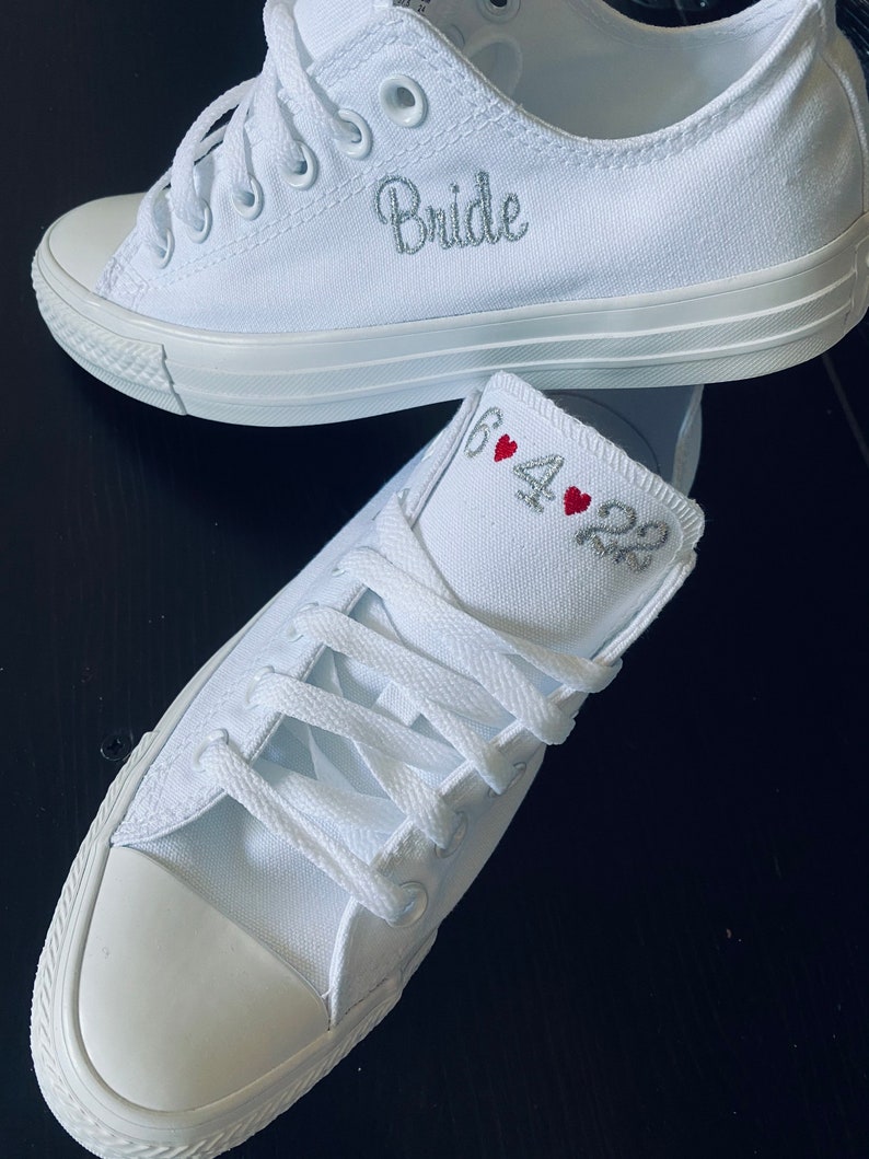 K&ouml;nnte beinhalten: Ein Paar wei&szlig;e Canvas-Sneaker mit dem Wort "Bride" auf einem Schuh und dem Datum "6.4.22" mit roten Herzen auf dem anderen Schuh bestickt.