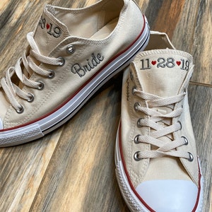 K&ouml;nnte beinhalten: Ein Paar cremefarbene Canvas-Sneaker mit rotem Besatz. Ein Schuh hat das Wort "Bride" bestickt, der andere das Datum "11.28.19".